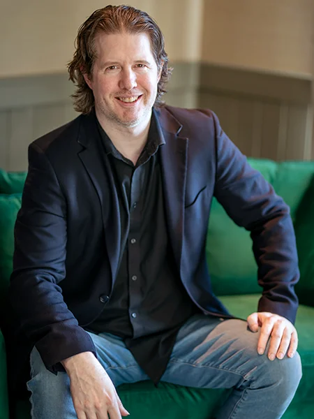 james ritzman - a white male wearing a blue blazer over a black button-up shirt and dress jeans - sitting on a couch in a conference room, smiling